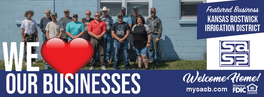 The image features a group of people standing together in front of a blue building. The individuals are of varying heights and many are wearing hats. In front of the group, there is a large heart icon with the words “WE ❤️ OUR BUSINESSES” written above it. On the top right of the image, there is text reading "Featured Business Kansas Bostwick Irrigation District". To the right, there is a "Welcome Home!" sign, below which, the words "mysasb.com" and "FDIC" are written. The overall sentiment of the image conveys positive vibes and appears to celebrate local businesses.