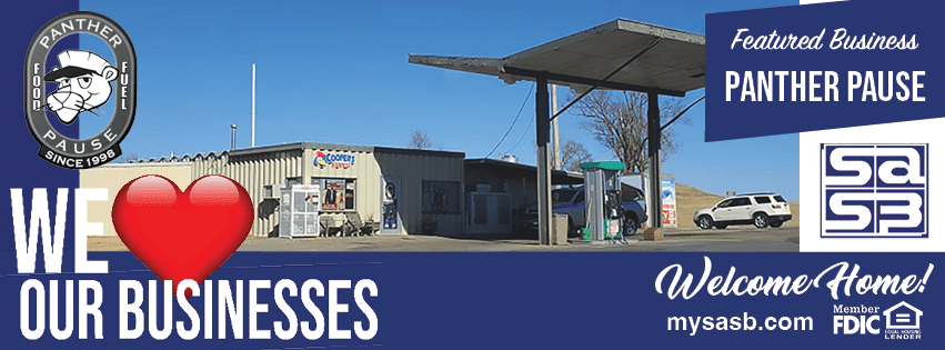 The image depicts a gas station with several parked vehicles including a white SUV. The gas station features two distinct blue banners. The top banner reads "Featured Business Panther Pause," while the banner below reads "We Love Our Businesses," using a heart symbol for the word "Love." The banners highlight a campaign to support local businesses. The bottom part of the image includes more text in the form of "Welcome Home!" and a website reference - "mysasb.com."